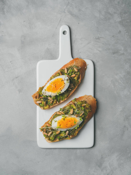 Top view of whole-grain toasts plated avocado with eggs, fresh mini herbs, sunflower seeds and sprinkled black salt served on ceramic white board - Fotó, kép