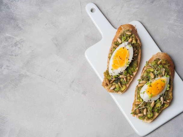 Fresh whole-grain toasts plated avocado with eggs, fresh mini herbs, sunflower seeds and sprinkled black salt served on ceramic white board - Foto, Imagem