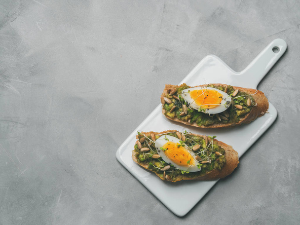 Fresh whole-grain toasts plated avocado with eggs, fresh mini herbs, sunflower seeds and sprinkled black salt served on ceramic white board - Foto, afbeelding