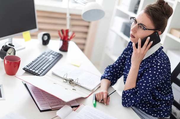 Uma jovem de óculos senta-se no escritório em uma mesa de computador e está falando ao telefone. Antes dela na mesa estão pastas com documentos
. - Foto, Imagem