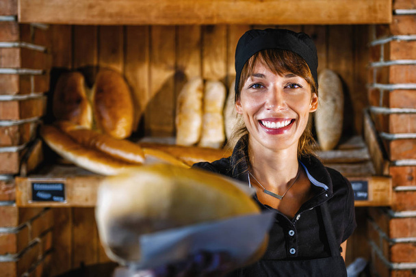Красивая улыбающаяся помощница пекаря, работающая в пекарне. Ношение черной униформы и продажа свежего хлеба
.   - Фото, изображение