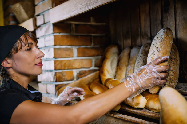 Mooie glimlachend bakkerij assistent dragen zwart uniform en het organiseren van brood in bakkerij winkel.  - Foto, afbeelding