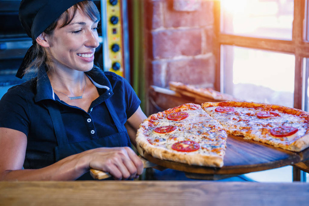 Mulher sorridente chef vestido de uniforme preto segurando casca de madeira com pizza no restaurante pizza
. - Foto, Imagem