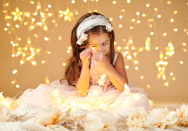 Gyermek lány játszik a karácsonyi fények, sárga háttér, öltözve rózsaszín ruhát. Ő keres a csillag fény. - Fotó, kép