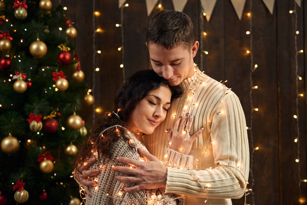young couple is in christmas decoration and lights, fir tree on dark wooden background, new year holiday concept - Photo, Image