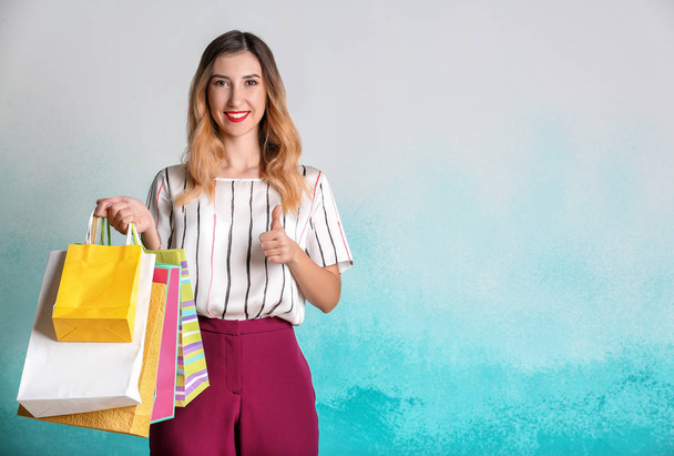 Hermosa mujer joven con bolsas de compras que muestran el pulgar hacia arriba en el fondo de color
 - Foto, Imagen