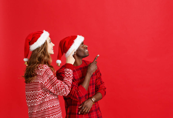 ευτυχισμένο ζευγάρι χαρούμενα μαύρο άνδρα και καυκάσιος γυναίκα σε Χριστούγεννα καπέλα στο κόκκινο backgroun - Φωτογραφία, εικόνα