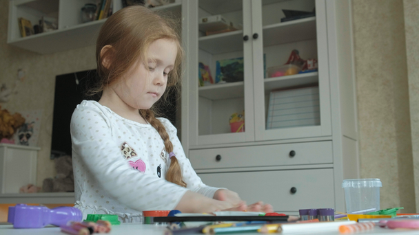 uma menina brinca com a plasticina, rola com as mãos, há figuras e lápis coloridos na área de trabalho, o desenvolvimento de habilidades motoras finas
 - Filmagem, Vídeo