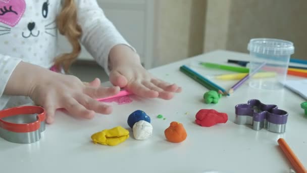 a little girl plays with plasticine, rolls it with her hands, there are figures and colorful pencils on the desktop, the development of fine motor skills - Footage, Video
