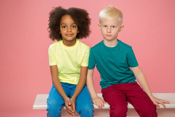 Side view of little kids. Happy couple caucasian and mixed race children are sitting nearby. Isolated on a pink background.  - Valokuva, kuva