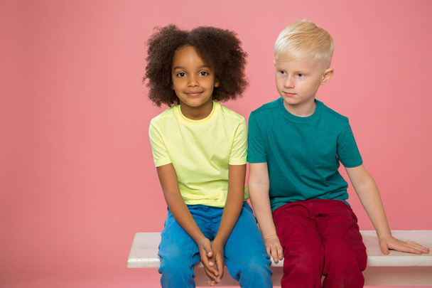 Seitenansicht von kleinen Kindern. Glückliches Paar kaukasische und gemischte Rassenkinder sitzen in der Nähe. Vereinzelt auf rosa Hintergrund.  - Foto, Bild