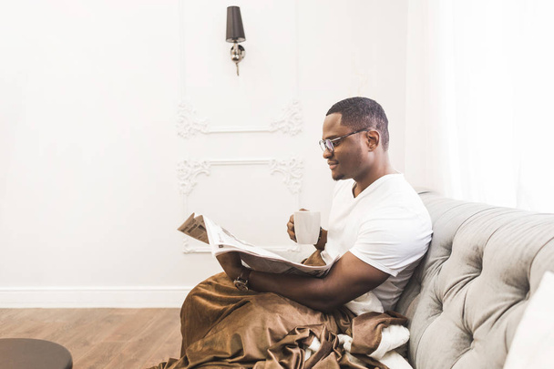 Young African American man, covered with a blanket reading a newspaper at home. - 写真・画像