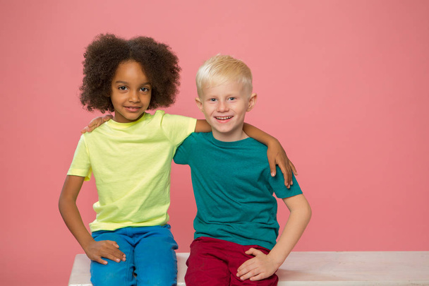 Vista laterale dei bambini piccoli. Buona coppia caucasica e africana bambini americani stanno sorridendo, seduto nelle vicinanze. Isolato su sfondo rosa. contatto con gli occhi. - Foto, immagini
