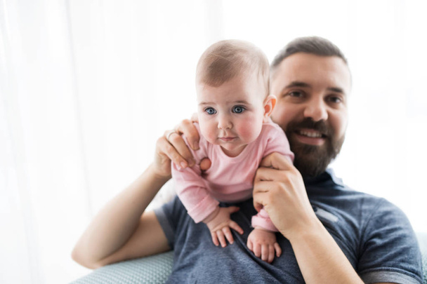 Portrait of young father with baby daughter sitting indoors on a sofa. - Foto, afbeelding