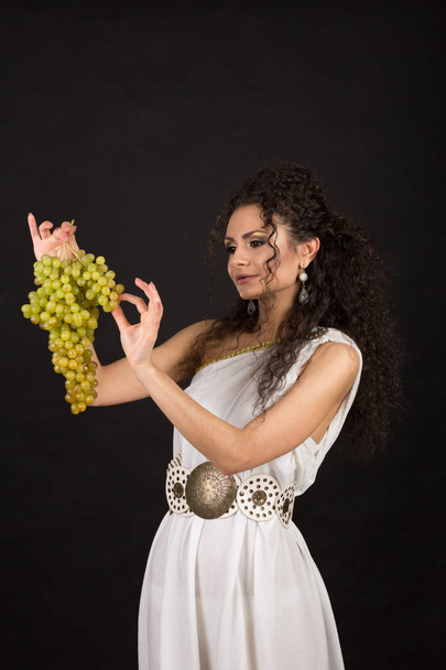 Portrait of a pretty young brunette in white greece toga holding a bunch of grapes - Photo, image