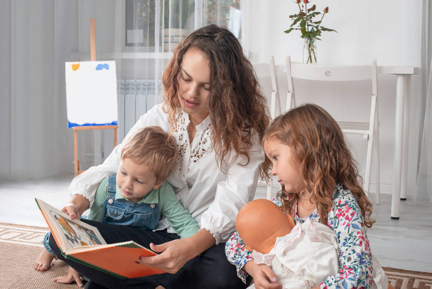 Joven madre o niñera con niños pequeños, un niño y una niña, sentarse en el suelo en una alfombra en la habitación en casa y leer un libro
 - Foto, imagen