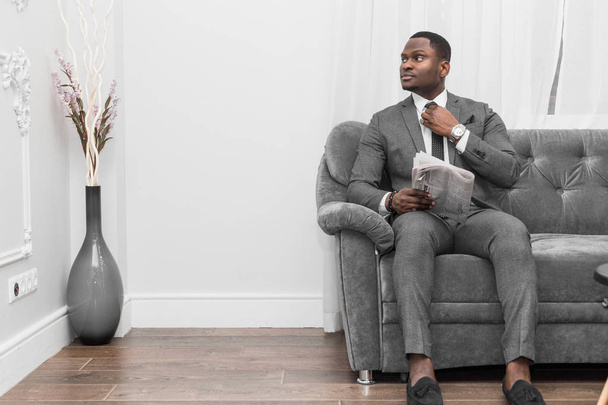 Young African American businessman in a gray suit reading a newspaper while sitting on a sofa. - Fotó, kép