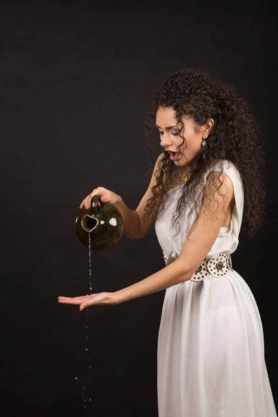 Curly girl in a Greek toga pouring water from a jug - Foto, Imagem