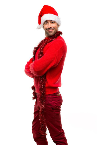 Man with red clothes celebrating the Christmas holidays keeping the arms crossed in lateral position while smiling on isolated white background - Photo, Image