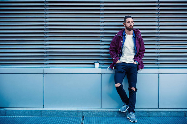 Красивый молодой человек в фиолетовой зимней куртке пьет кофе. Парень, пьющий кофе на улице
. - Фото, изображение