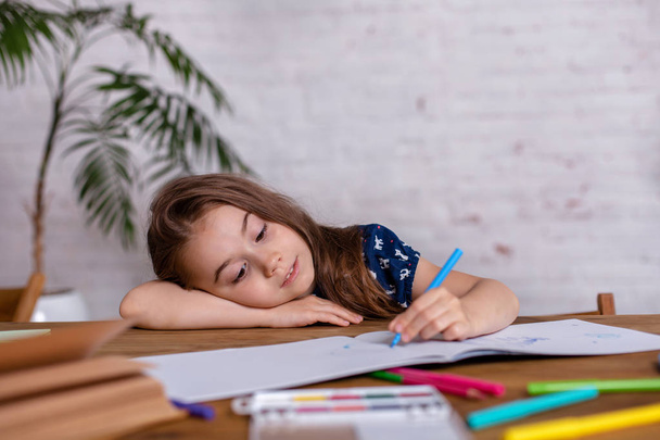inspiriertes kleines Mädchen am Tisch zeichnet mit Buntstiften oder erledigt Hausarbeit - Foto, Bild