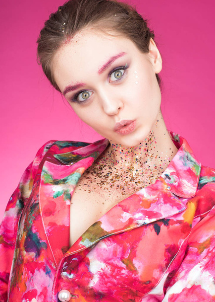 Colorful Studio portrait of a beautiful young woman with eyes wide open. Makeup with glitter. Depict and gesticulating hands. Pink background - Φωτογραφία, εικόνα