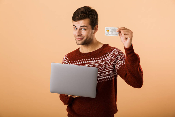 Portrait a smiling young man dressed in sweater isolated over beige background, holding laptop computer, showing credit card - Фото, изображение