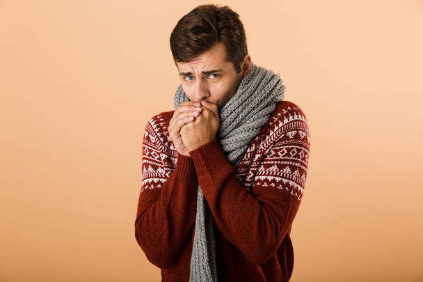 Portrait a young man dressed in sweater and scarf isolated over beige background, warming up arms - Photo, Image