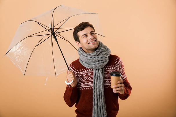 Portrait a happy young man dressed in sweater and scarf isolated over beige background, standing under umbrella, holding coffee cup, looking up - Photo, Image