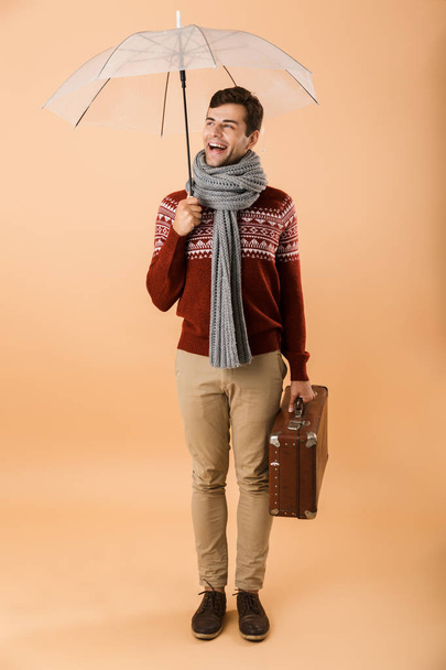 Full length portrait a smiling young man dressed in sweater and scarf isolated over beige background, standing under an umbrella, carrying briefcase - Fotoğraf, Görsel
