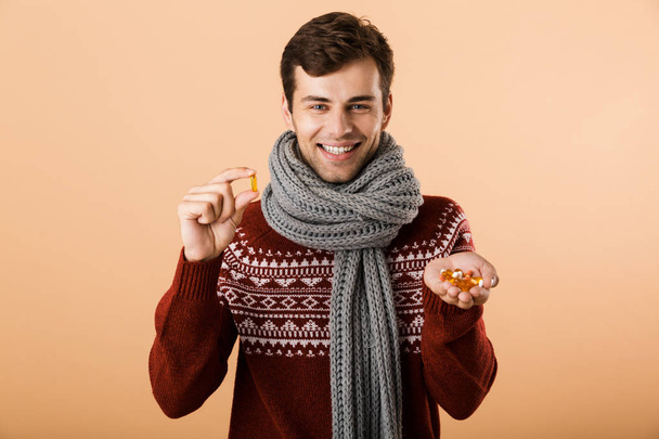 Portrait an excited man dressed in sweater and scarf isolated over beige background, showing pills capsules - Photo, image