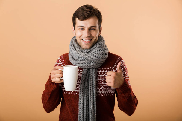 Portrait a cheerful man dressed in sweater and scarf isolated over beige background, holding cup of hot tea, showing thumbs up - Photo, image