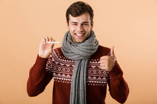 Portrait of a happy young man dressed in sweater and scarf isolated over beige background, showing thermometer, showing thumbs up - Zdjęcie, obraz