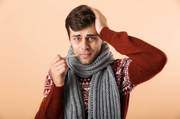 Portrait of a sick young man dressed in sweater and scarf isolated over beige background, showing thermometer - Foto, immagini