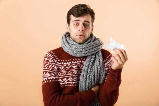 Πορτρέτο του ένα άρρωστο νεαρός άνδρας ντυμένος με πουλόβερ και κασκόλ απομονωμένη μπεζ φόντο, κρατώντας χαρτί ιστών - Φωτογραφία, εικόνα