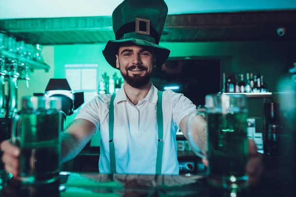 Saint Patrick's Day Party. Barman is Holding a Traditional Green Beer. Barman is Young Smiling Beard Man. Man Standing at Bar Counter. Man Wearing a Green Hat. Pub Interior. Green Tone. - Foto, imagen