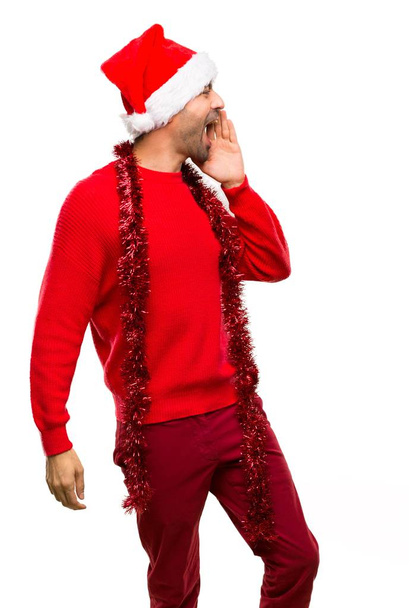 Man with red clothes celebrating the Christmas holidays shouting with mouth wide open to the lateral on isolated white background - Photo, Image