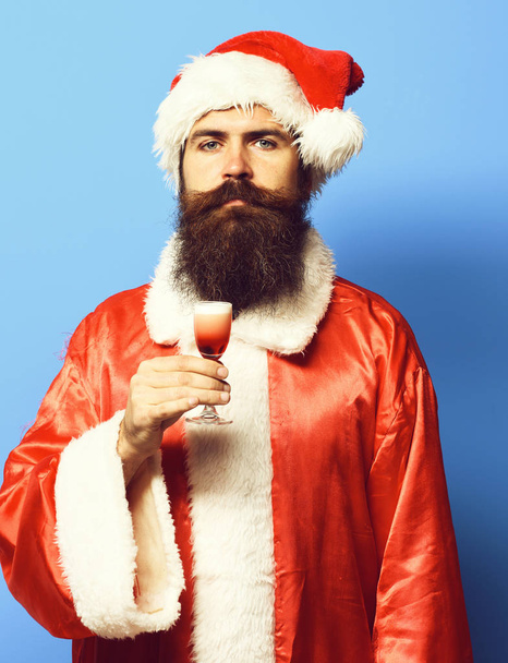 handsome bearded santa claus man with long beard on serious face holding glass of alcoholic shot in red christmas or xmas sweater and new year hat on blue studio background - Foto, afbeelding