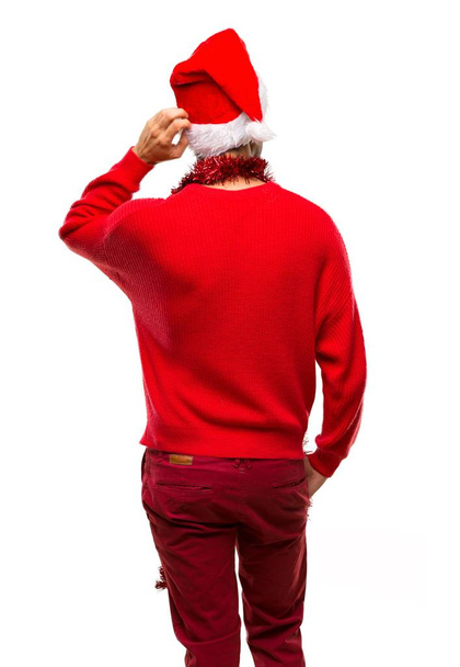 Hombre con ropa roja celebrando las vacaciones de Navidad en la posición trasera mirando hacia atrás mientras se rasca la cabeza sobre un fondo blanco aislado
 - Foto, Imagen