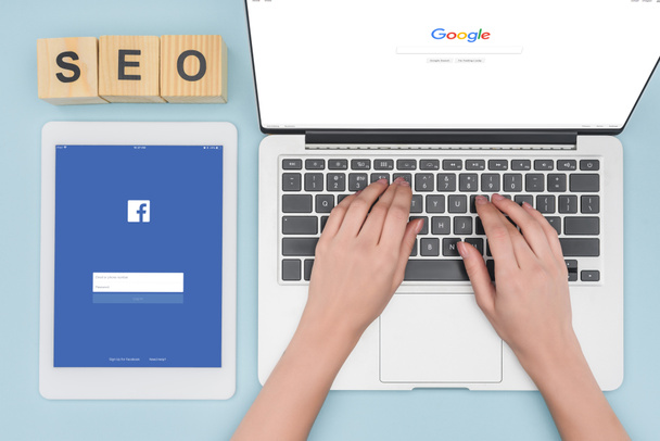 Вид сверху женщины, сидящей на клавиатуре ноутбука с открытым браузером Google рядом с цифровым планшетом с приложением Facebook на светло-голубом фоне
  - Фото, изображение