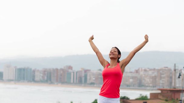 Successful sporty woman raising arms  towards city scape at Gijon, Asturias, Spain. Female fitness motivation and lifestyle concept. - Foto, Imagem