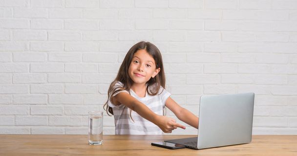 Nuori latino lapsi istuu pöydällä tietokoneen kannettavan erittäin onnellinen osoittaa käsin ja sormella
 - Valokuva, kuva