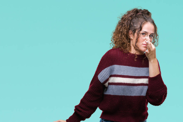 Beautiful brunette curly hair young girl wearing glasses over isolated background smelling something stinky and disgusting, intolerable smell, holding breath with fingers on nose. Bad smells concept. - Photo, Image