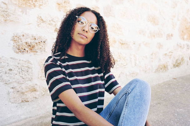 Gözlük, kot pantolon ve zemin ve kıvırcık saç üzerinde oturan bir çizgili tişört giyen ciddi genç siyah kadın portresi - Fotoğraf, Görsel