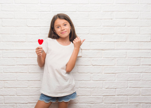 Jonge Latino jongen over witte bakstenen muur eten rood hart lollipop snoep aan te wijzen en te tonen met de duim aan de kant met een blij gezicht lachend - Foto, afbeelding