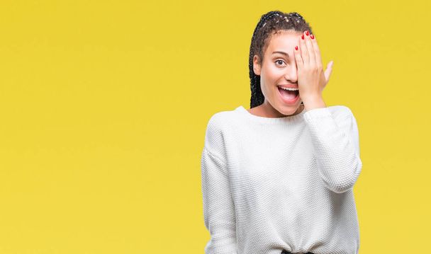 Молодий плетені волосся афро-американських дівчата носити взимку светр над ізольованих фон охоплюють одне око рукою з впевнено посмішки на обличчя і сюрприз емоцій. - Фото, зображення