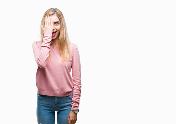 Giovane bella donna bionda indossa maglione invernale rosa su sfondo isolato che copre un occhio con mano con sorriso fiducioso sul viso e sorpresa emozione
. - Foto, immagini