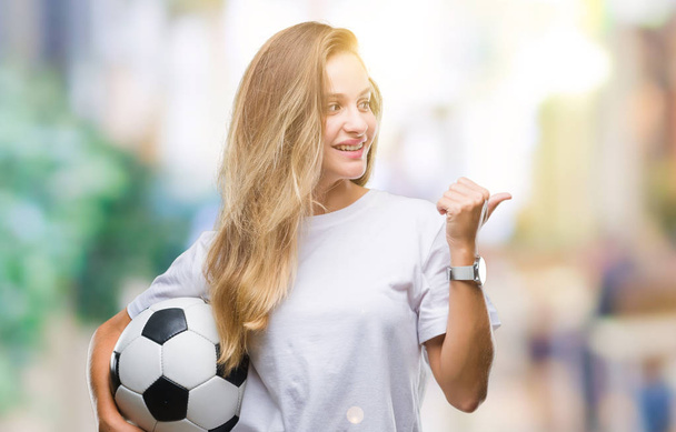 Jovem bela mulher loira segurando bola de futebol sobre fundo isolado apontando e mostrando com o polegar até o lado com o rosto feliz sorrindo
 - Foto, Imagem