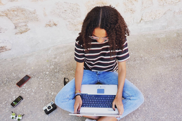 Een portret van gerichte jonge zwarte vrouw met krullend haar het dragen van bril, jeans en een gestreept t-shirt, naast technologie zoals smartphones en camera, zittend op de grond en werken of huiswerk maken - Foto, afbeelding