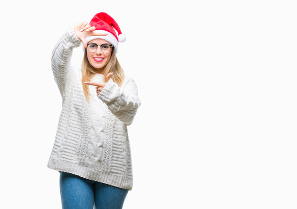 Молодая красивая женщина в рождественской шляпе на изолированном фоне улыбается делая рамку с руками и пальцами с счастливым лицом. Концепция творчества и фотографии
. - Фото, изображение